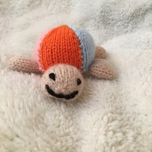 May include: A knitted toy turtle with a multi-colored shell. The shell is orange, pink, and blue. The turtle has a smiling face.
