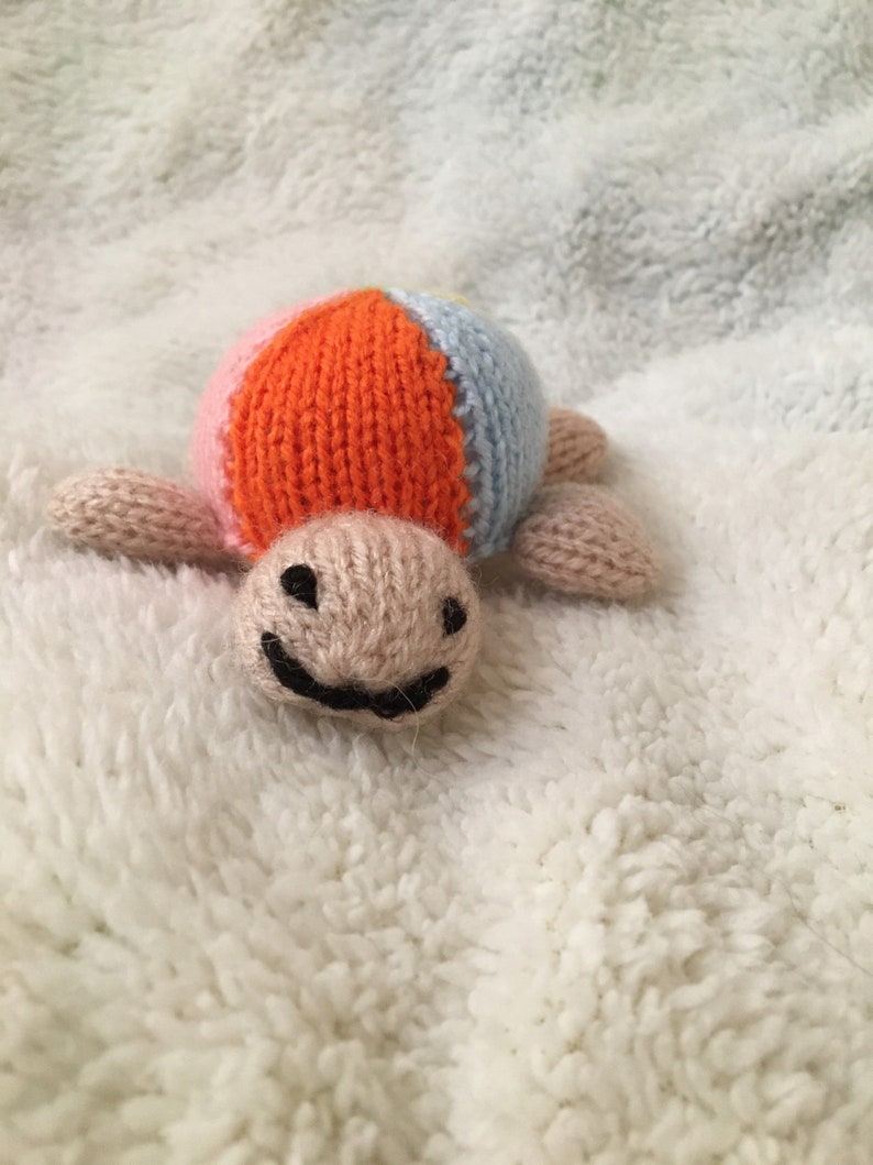 May include: A knitted toy turtle with a multi-colored shell. The shell is orange, pink, and blue. The turtle has a smiling face.