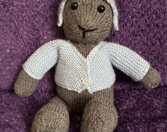May include: Hand-knitted grey stuffed toy wearing a light blue cardigan and a white hat with pink ears. The plush toy has black eyes and is sitting on a purple textured surface. The toy is a handmade item.