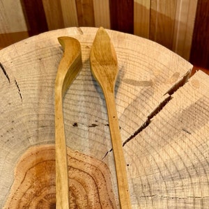 May include: Two wooden spoons, one with a curved handle and one with a straight handle, are displayed on a wooden surface.
