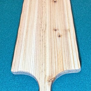 May include: A long, rectangular wooden serving board with a tapered handle featuring a circular hole at the end. The board displays a light tan color with prominent vertical wood grain patterns and several small, dark brown knots throughout the surface.