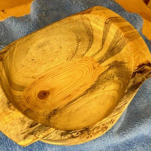 May include: A hand-carved wooden bowl featuring a natural light tan finish with prominent dark brown grain patterns and knots. The bowl has a thick, irregular rim and a deep, rounded interior, showcasing the organic texture of the wood grain.