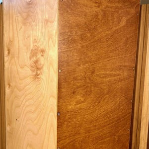 Könnte beinhalten: Eine rechteckige Cajon-Trommel aus Holz mit einer hellen Frontplatte aus Naturholz mit sichtbarer Maserung und einer dunkleren, braun gebeizten Seitenwand. Die Seitenwand ist mit mehreren kleinen Metallschrauben am Rahmen befestigt.