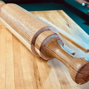 May include: A wooden rolling pin with a light brown barrel and darker brown handles. The rolling pin is made of wood and has a smooth, polished finish. It is designed for rolling out dough for baking.