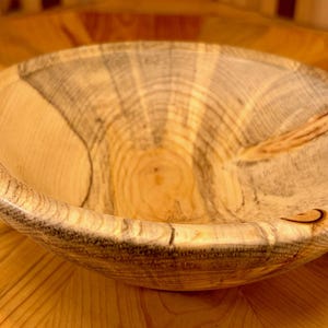 May include: A round wooden bowl featuring a light tan and cream color palette with prominent dark grey grain patterns. The bowl displays a smooth, polished interior surface with visible natural wood knots and organic textures along the rim.