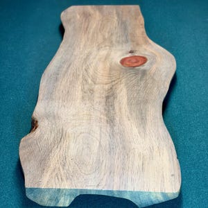 May include: A rectangular wooden serving board with a natural live edge, featuring a prominent reddish-brown knot in the center. The wood grain displays light and dark brown swirls throughout the surface, showcasing a smooth, flat top texture.