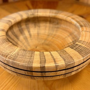 May include: A round wooden bowl featuring a light tan and dark gray striped grain pattern. The bowl has a wide, flat rim and a deep, smooth interior basin. Two thin, dark horizontal lines are carved into the exterior side of the bowl.