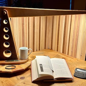 May include: A wooden desk lamp with a unique design, featuring a triangular base with circular cutouts. The lamp is illuminated and casts a warm glow on a book, a mug, and a calculator on a wooden desk.