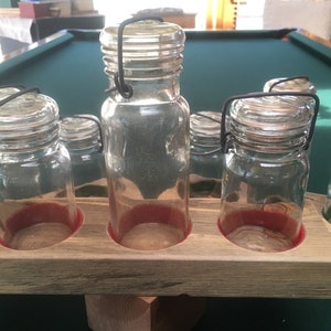 Puede incluir: Un estante de madera con cinco tarros de vidrio con asas de alambre. Los tarros son transparentes y tienen un borde rojo. El estante está sobre una superficie de fieltro verde.