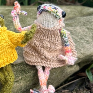 May include: Two knitted frog toys wearing sweaters. One frog is wearing a yellow sweater and the other is wearing a brown sweater with a colorful scarf. The frogs are sitting on a rock.