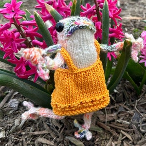 May include: A knitted frog doll wearing a yellow dress with a white crocheted bodice and a floral pattern on its body. The frog is standing in a bed of pink hyacinths.