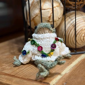May include: A handmade, knitted frog figurine wearing a white sweater adorned with colorful sequin accents. The frog has green and gray knitted details, black eyes, and is posed on a wooden surface. The background includes decorative spheres within a black wire basket.