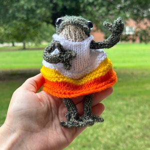 May include: A knitted frog wearing a white and orange candy corn costume. The frog is holding its right arm up in the air.