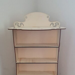 Può includere: Un'unità a scaffale in legno con quattro ripiani e decorazioni a volute in alto e in basso. Lo scaffale è in legno chiaro e ha una finitura naturale.