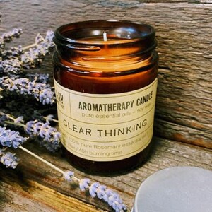 May include: A brown glass jar candle with a white label that reads "AROMATHERAPY CANDLE pure essential oils + soy wax" and "CLEAR THINKING 100% pure Rosemary essential oil Soy wax | 40h burning time".