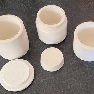 May include: Five white ceramic containers with lids. The containers are cylindrical and have a smooth, matte finish. The lids are round and fit snugly on top of the containers.