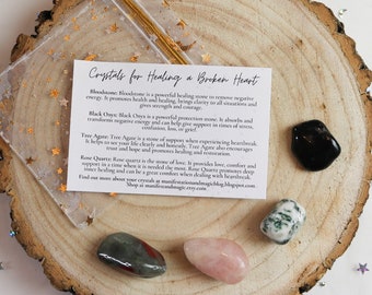 May include: A set of five crystals for healing a broken heart. The crystals are bloodstone, black onyx, tree agate, rose quartz, and a green and red crystal. The crystals are arranged on a wooden surface with a white card that reads "Crystals for Healing a Broken Heart".