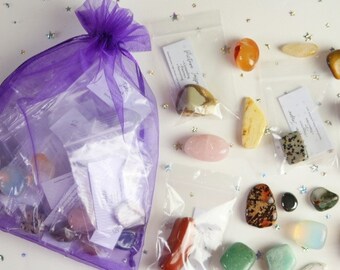 Op de afbeelding: Een paarse gaas tas gevuld met verschillende getrommelde stenen en kristallen. De stenen zitten in individuele doorzichtige plastic zakjes met kleine witte kaartjes. De stenen omvatten rozenkwarts, amethist, helder kwarts en meer.