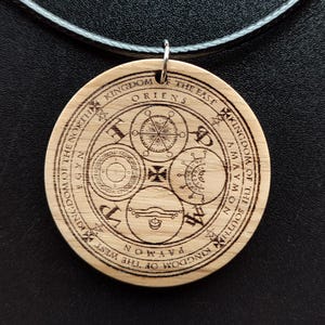 Puede incluir: Un colgante redondo de madera con símbolos y texto grabados, suspendido de un cordón negro. El colgante presenta diseños intrincados dentro de un marco circular, con texto como "Reino del Este" y "Reino del Oeste".