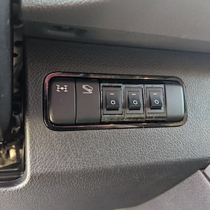 May include: A close-up of a black panel with three toggle switches and two buttons. The buttons have icons depicting a trailer and a car with a downward arrow. The switches are labelled with a minus sign.
