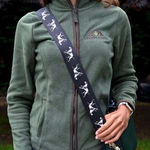 May include: A green fleece jacket with a brown trim and a black strap with white dog silhouettes. The jacket has a zipper closure and a small logo on the chest. The person is wearing blue jeans and a dark green purse.