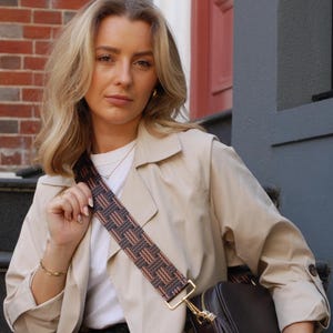 May include: A dark brown leather crossbody bag with a decorative tassel and gold-tone hardware. The bag features a patterned strap in shades of brown and tan. The person is wearing a beige trench coat and a white shirt.