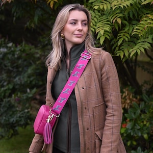 May include: A pink crossbody bag with a matching strap featuring a repeating dog pattern. The bag has a tassel and gold-colored hardware. The woman is wearing a brown plaid jacket and a green zip-up jacket.