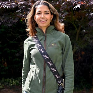 May include: An olive green fleece jacket with a brown zipper and trim. A black strap with a dog pattern is worn across the body. The jacket has a small embroidered logo on the chest and is paired with blue jeans.