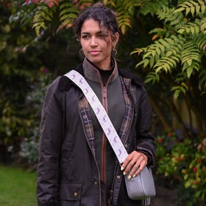 May include: A woman wearing a dark brown jacket with a gray crossbody bag. The bag strap is white with a repeating purple dog illustration. The jacket has a corduroy collar and a plaid lining. The background is a green garden.