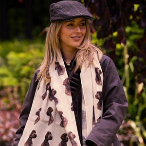 May include: A cream scarf with a repeating pattern of brown dog illustrations. The scarf is worn with a dark jacket and a brown tweed flat cap.