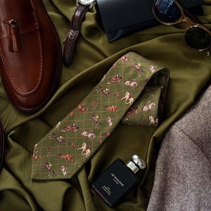May include: An olive green tie featuring a horse rider pattern, a brown leather watch, brown leather loafers, sunglasses, a black wallet, and cologne are arranged on a green satin surface.