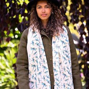 May include: A cream-colored scarf with a blue dragonfly pattern is draped over a green jacket. The scarf features a repeating design of dragonflies in shades of blue and brown. The jacket has a plaid pattern and is buttoned up.