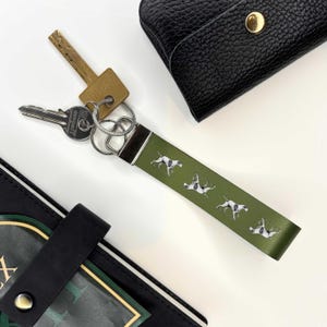May include: Green leather keyring with a silver key ring and four white and black dog illustrations. The keyring is attached to a set of keys.