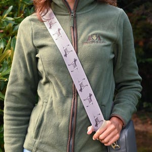 Puede incluir: Una correa de hombro gris claro con un patrón repetido de ilustraciones de perros. La correa se lleva sobre una chaqueta de forro polar verde. La chaqueta tiene una cremallera y ribete marrones. Se ve un bolso negro.