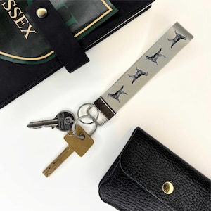 May include: A grey key chain with a silver metal ring and four black and white dog silhouettes. The key chain is attached to two keys.
