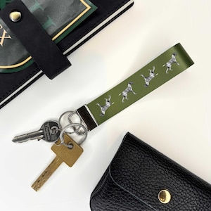 Può includere: Un portachiavi in ​​pelle verde con un anello portachiavi argentato e quattro silhouette di cani bianche. Il portachiavi è attaccato a un set di chiavi.