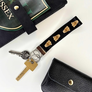 May include: A black key chain with a silver ring and four small brown dog faces printed on the strap. The key chain is attached to two keys, one gold and one silver.
