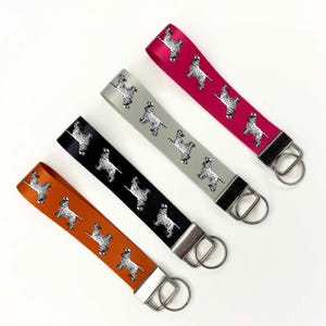 May include: Four keychains with different coloured ribbon straps and a silver key ring. Each strap features a repeating pattern of a black and white dog silhouette.