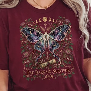 May include: A maroon short-sleeve t-shirt featuring a graphic of a large moth with celestial patterns on its wings, surrounded by floral elements and moon phases, with the text Fae Bargain Survivor printed in a gold-toned font at the bottom.
