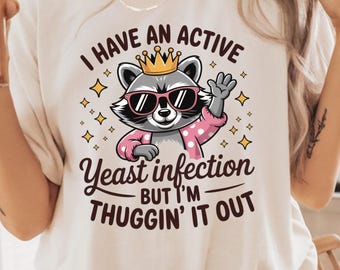 Könnte beinhalten: Ein weißes T-Shirt mit einem Aufdruck eines Waschbären, der eine Krone und eine Sonnenbrille trägt. Der Text auf dem Shirt lautet "Ich habe eine aktive Hefeinfektion, aber ich bin dabei, sie zu besiegen."