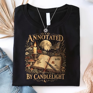 May include: A black t-shirt featuring a graphic print of an open book, a lit candle, a fountain pen, mushrooms, and a full moon. The text "ANNOTATED BY CANDLELIGHT" is printed in gold lettering above and below the central illustration.
