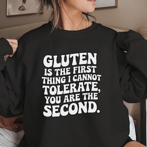 May include: Black sweatshirt with the white text: "GLUTEN IS THE FIRST THING I CANNOT TOLERATE, YOU ARE THE SECOND." The sweatshirt is a crew neck style. The image is a close-up of the upper body.