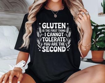 May include: Black t-shirt with a white graphic that reads "GLUTEN is the first thing I CANNOT TOLERATE YOU ARE THE SECOND." The design includes wheat stalks and stars. The shirt is worn by a person with blonde hair.