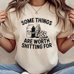 May include: Cream-colored t-shirt with the phrase "SOME THINGS ARE WORTH SHITTING FOR" in black. The graphic includes a milk carton, ice cream cone, and cheese. The shirt is short-sleeved.