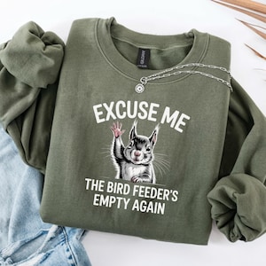 May include: An olive green sweatshirt with the text "EXCUSE ME" above an illustration of a squirrel waving. Below the squirrel is the text "THE BIRD FEEDER'S EMPTY AGAIN". A silver necklace is draped across the sweatshirt.