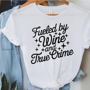 May include: White t-shirt with black text that reads "Fueled by Wine and True Crime" with stars around the text.