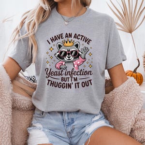 May include: Gray t-shirt with a cartoon raccoon wearing sunglasses and a crown. The text on the shirt reads "I have an active yeast infection but I'm thuggin' it out."