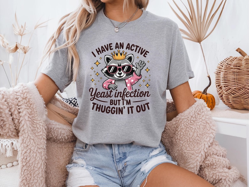 May include: Gray t-shirt with a cartoon raccoon wearing sunglasses and a crown. The text on the shirt reads "I have an active yeast infection but I'm thuggin' it out."