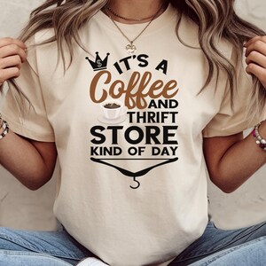 May include: A beige t-shirt with the text "It's a Coffee and Thrift Store Kind of Day" printed on it. The text is in black with a brown coffee cup and a crown graphic.
