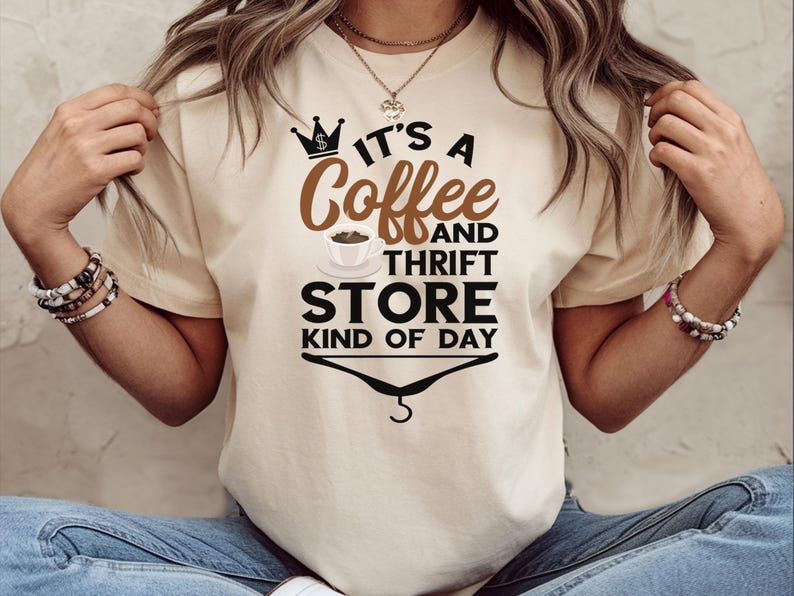 May include: A beige t-shirt with the text "It's a Coffee and Thrift Store Kind of Day" printed on it. The text is in black with a brown coffee cup and a crown graphic.